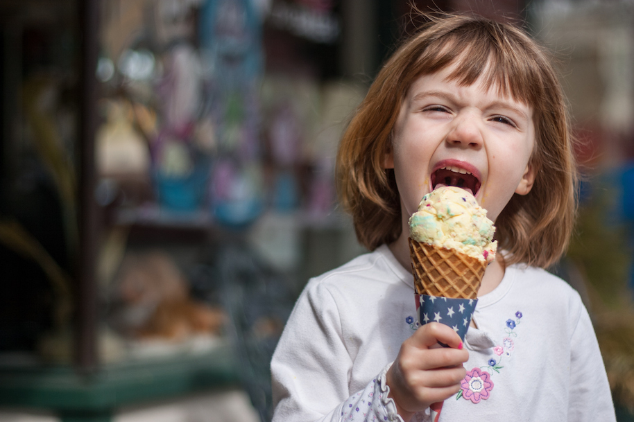 This is a close up color image of a cute little girl enjoying a waffle cone. She is casually dressed with an ice cream stand in the background.